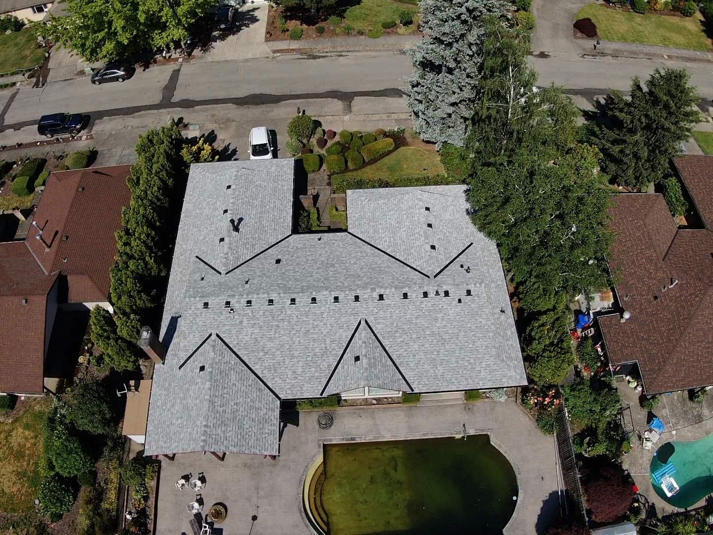 Aerial view of a large house with a gray roof, surrounded by trees and neighboring homes, featuring a swimming pool in the backyard.
