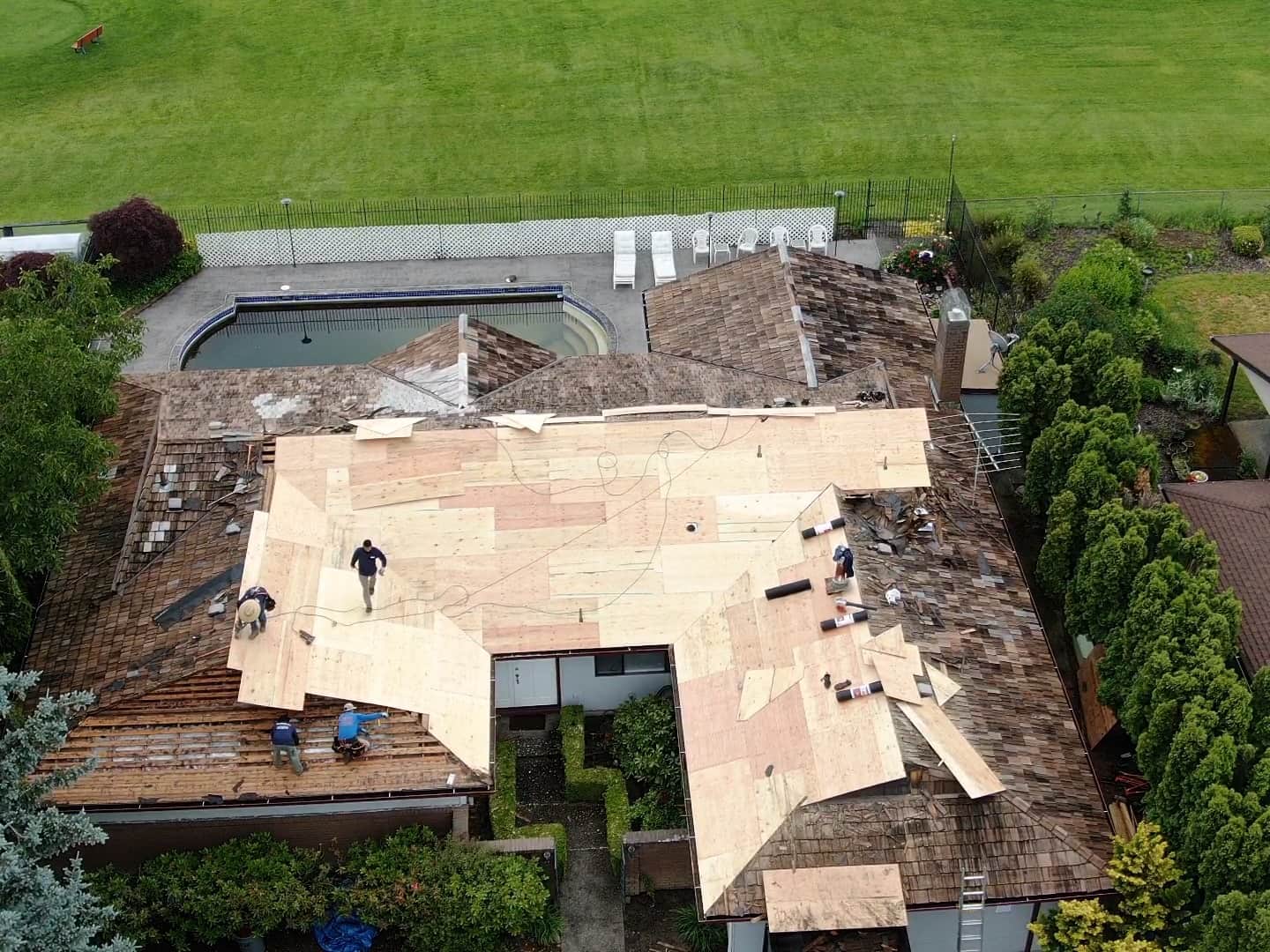 A house with a wooden roof undergoing renovation, with several workers installing plywood sheets. A swimming pool is visible in the backyard, surrounded by green grass.