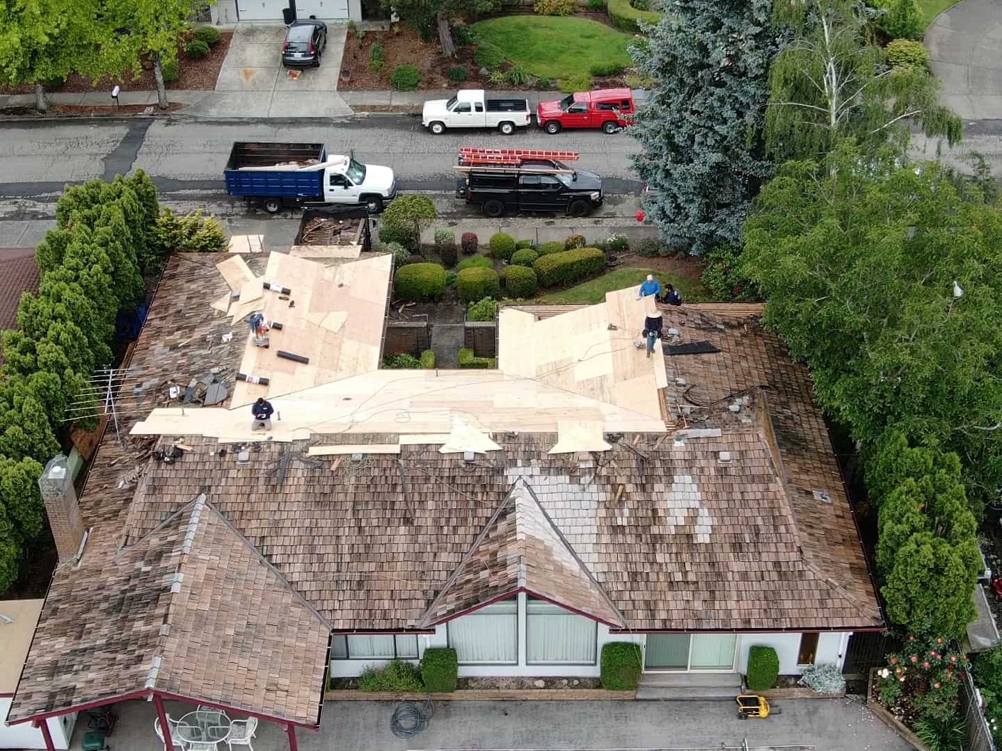 Aerial view of a house with workers replacing the roof. Wooden sheathing is visible, and construction materials are on the roof. Trees and a street with parked trucks surround the property.