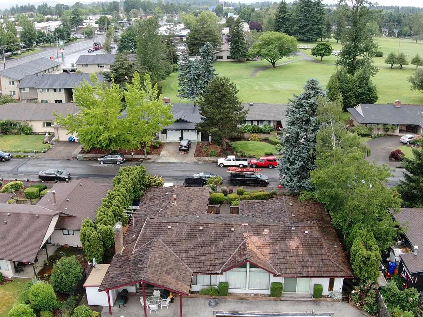 Aerial view of a residential neighborhood with several houses surrounded by trees, a road with parked vehicles, and a golf course in the background.